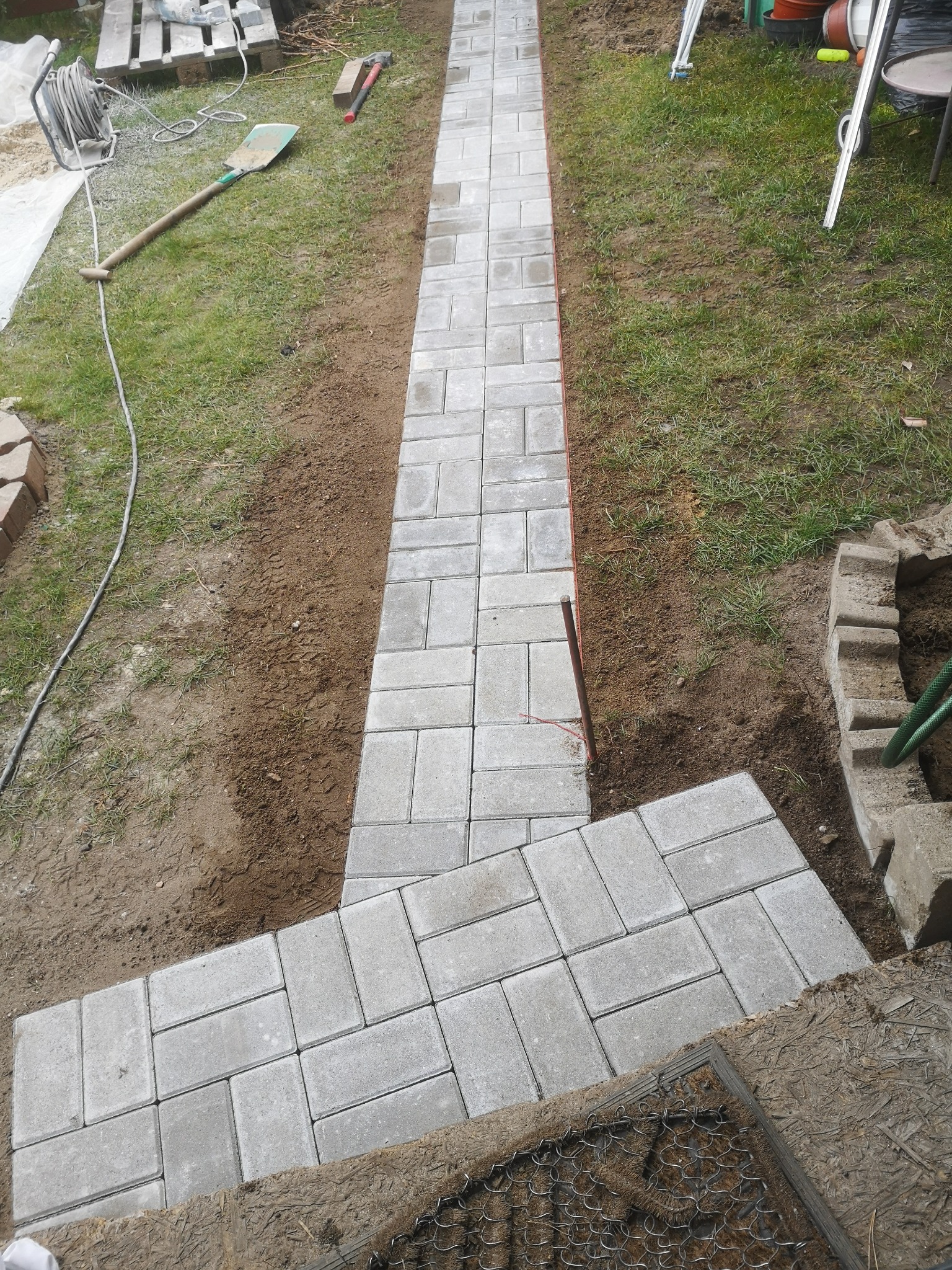 Świeżo ułożona ścieżka z szarej kostki brukowej o prostokątnym kształcie, prowadząca przez trawnik z widocznymi narzędziami: łopata, młotek i zwinięty kabel elektryczny.