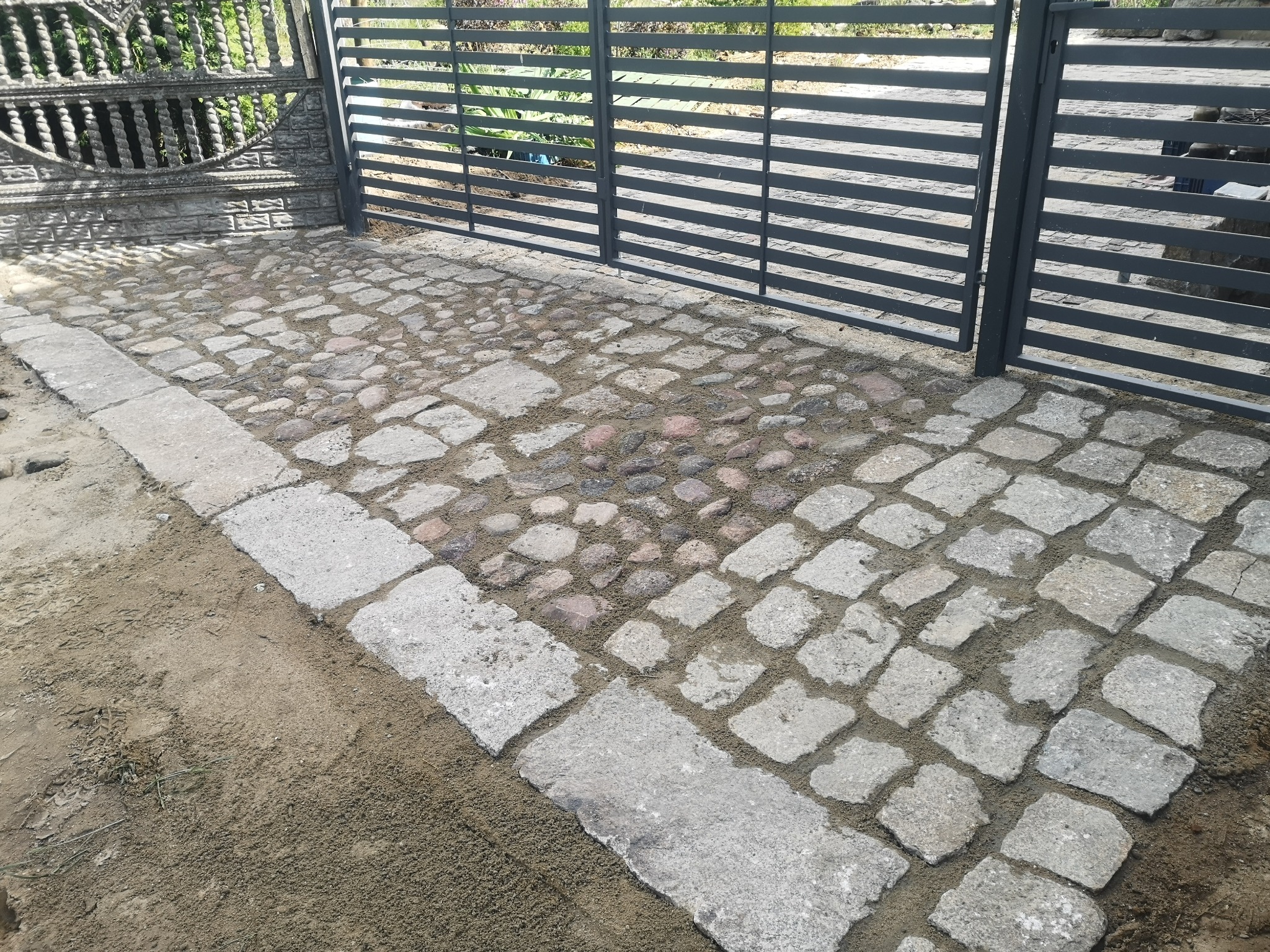 Ułożona kostka granitowa w odcieniach szarości i różu, obramowana betonowym krawężnikiem, widoczna brama wjazdowa z metalowymi przęsłami.