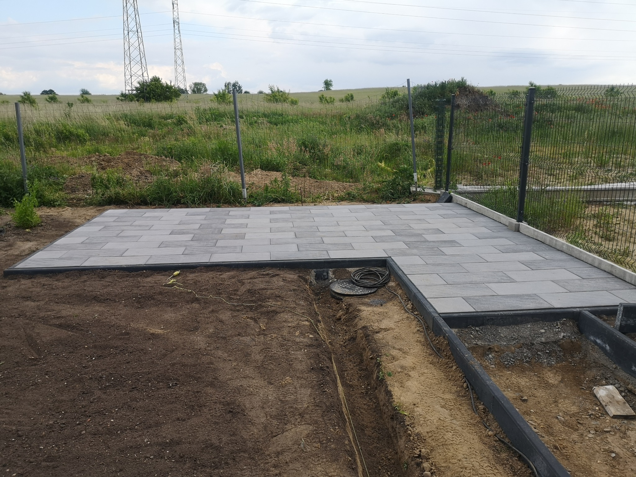 Układanie szarej kostki brukowej na częściowo przygotowanym podłożu, widoczne kable i wykop, w tle łąka i słupy wysokiego napięcia.