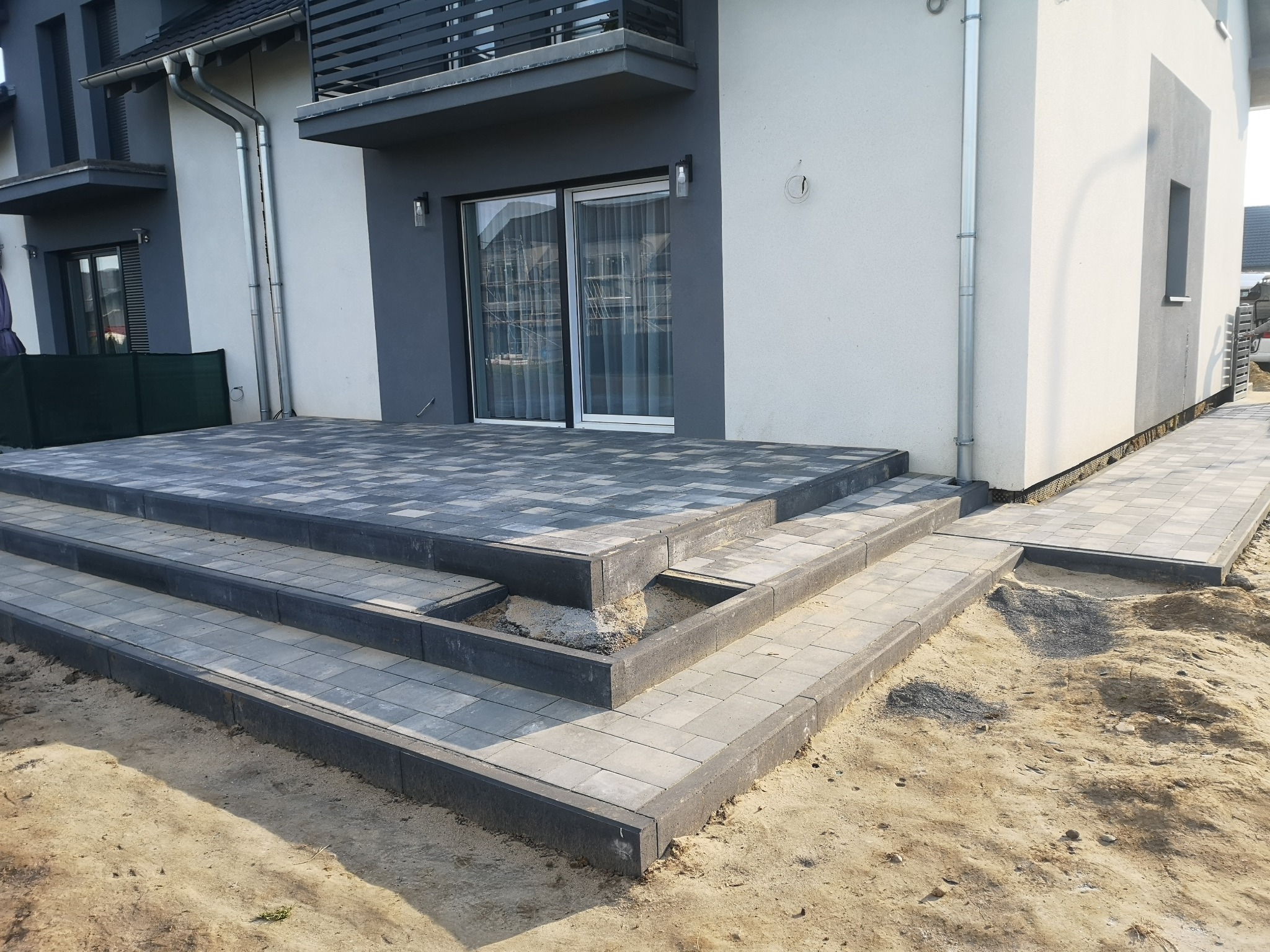 Nowoczesny taras z kostki brukowej w odcieniach szarości, prowadzący schodami do drzwi balkonowych, z widocznym fragmentem niedokończonej krawędzi i piaskowym podłożem.