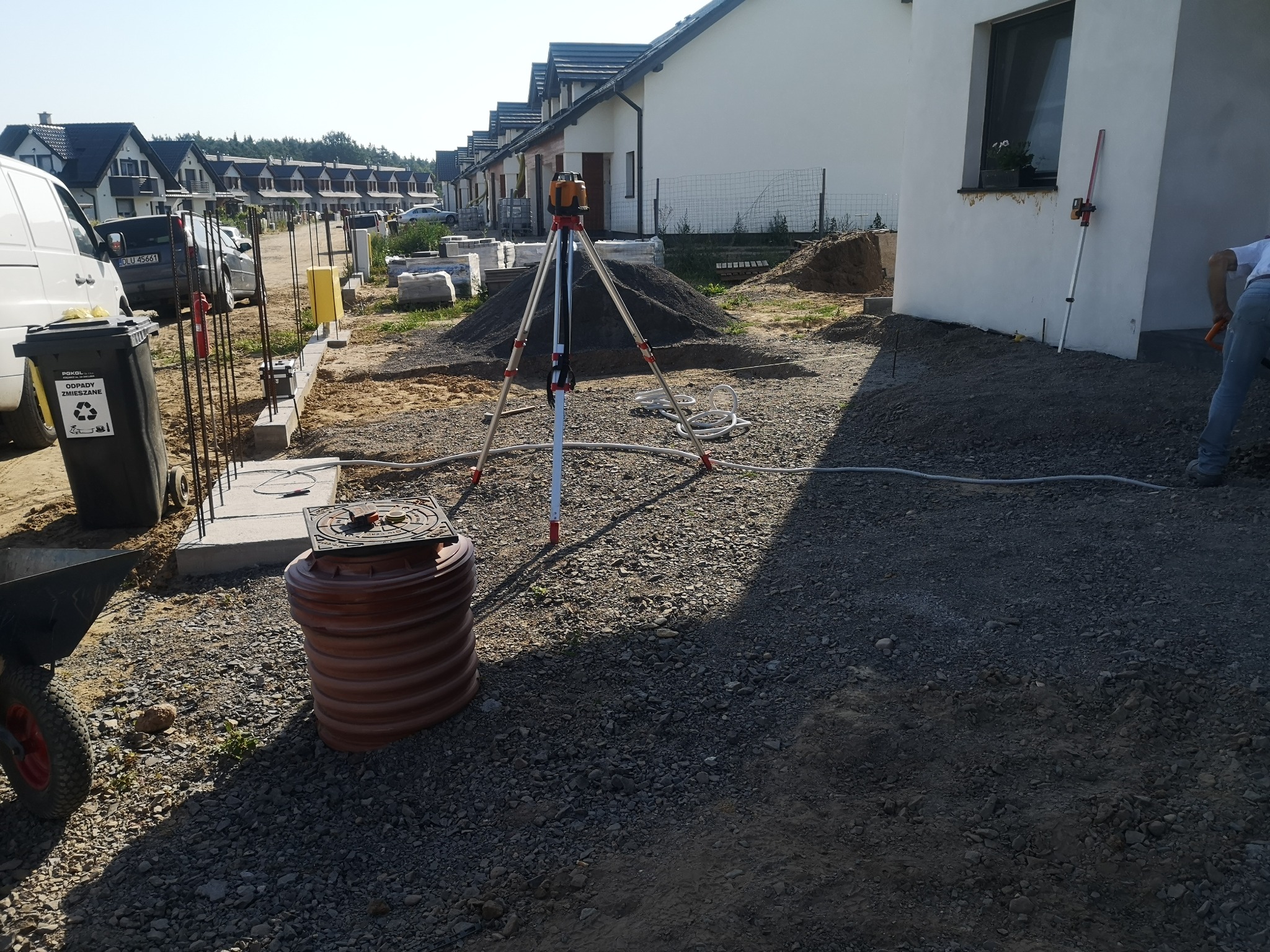 Teren budowy z niwelatorem laserowym na statywie, stosem rur z tworzywa sztucznego, taczką i fundamentami w tle szeregu domów jednorodzinnych.
