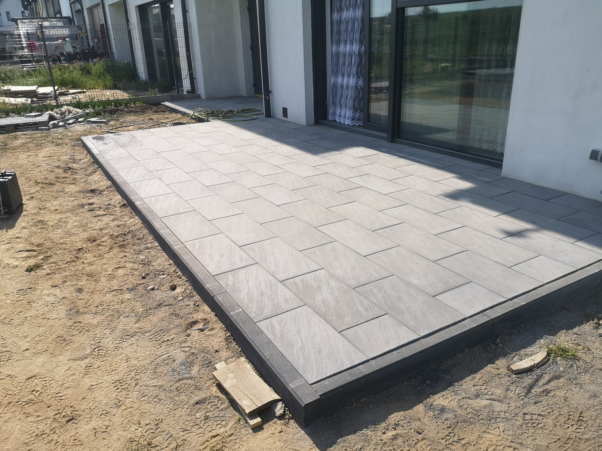 Nowoczesny taras z szarych płyt betonowych ułożonych na piasku przy białym budynku z dużymi oknami przesuwnymi.