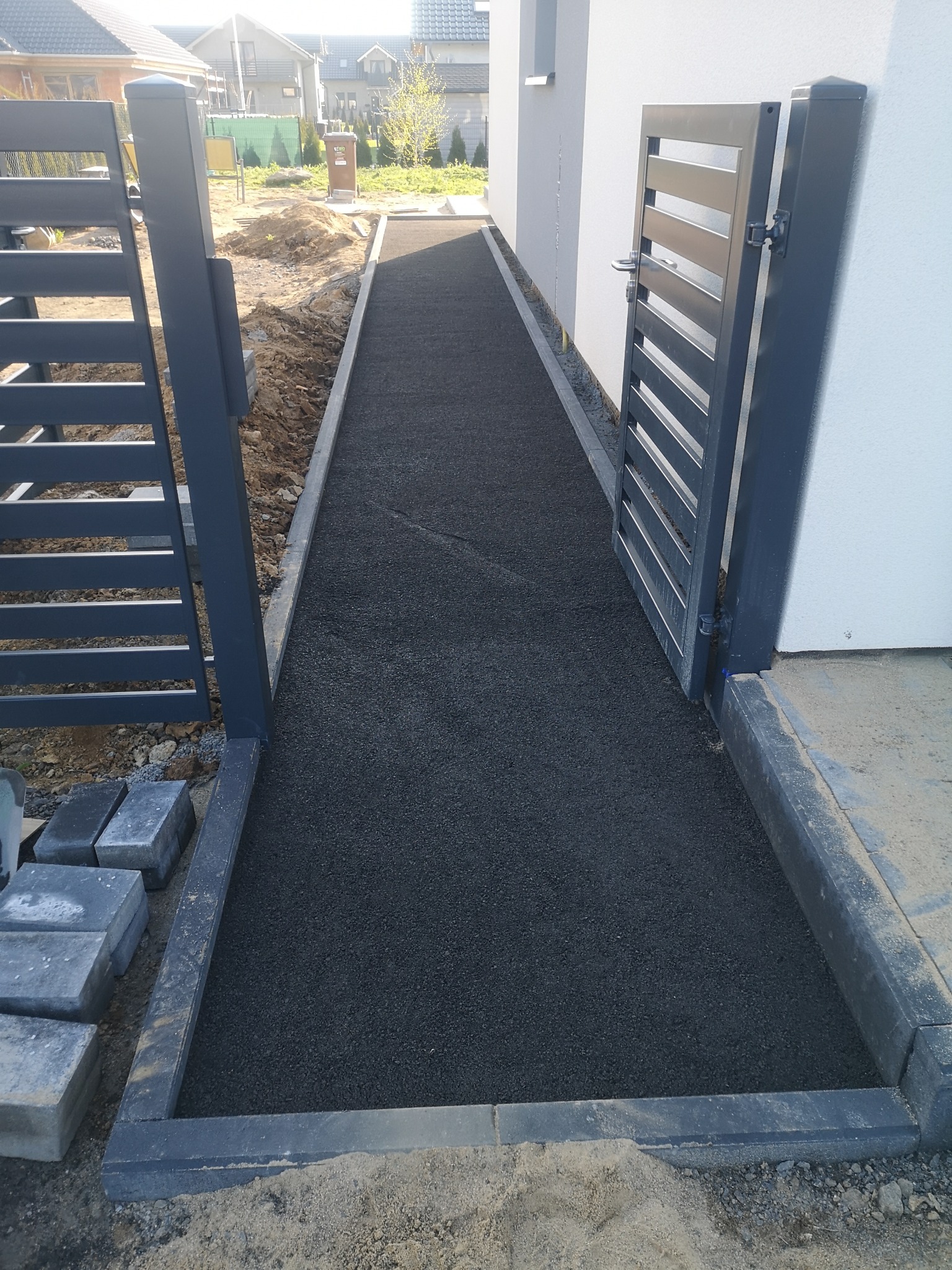 Ścieżka z drobnego, ciemnego kruszywa prowadząca od furtki do wejścia domu, obramowana szarą kostką i widoczne niedokończone prace ziemne po bokach.