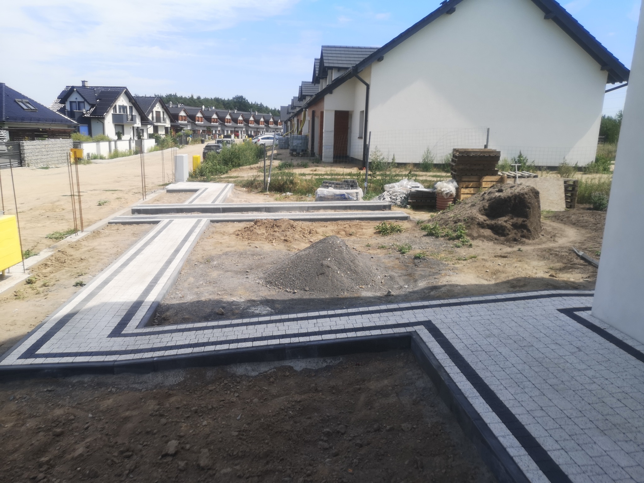 Nowo ułożona, szara kostka brukowa z ciemnoszarą obwódką tworząca ścieżkę wzdłuż szeregu domów jednorodzinnych w trakcie budowy, widoczna sterta piasku i palety z kostką.