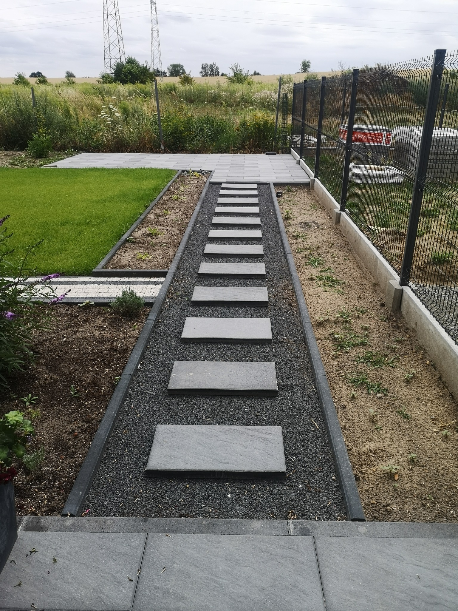 Ścieżka z szarych płyt ułożona w czarnym żwirze, prowadząca przez ogród z trawnikiem i rabatami, ogrodzona siatką.