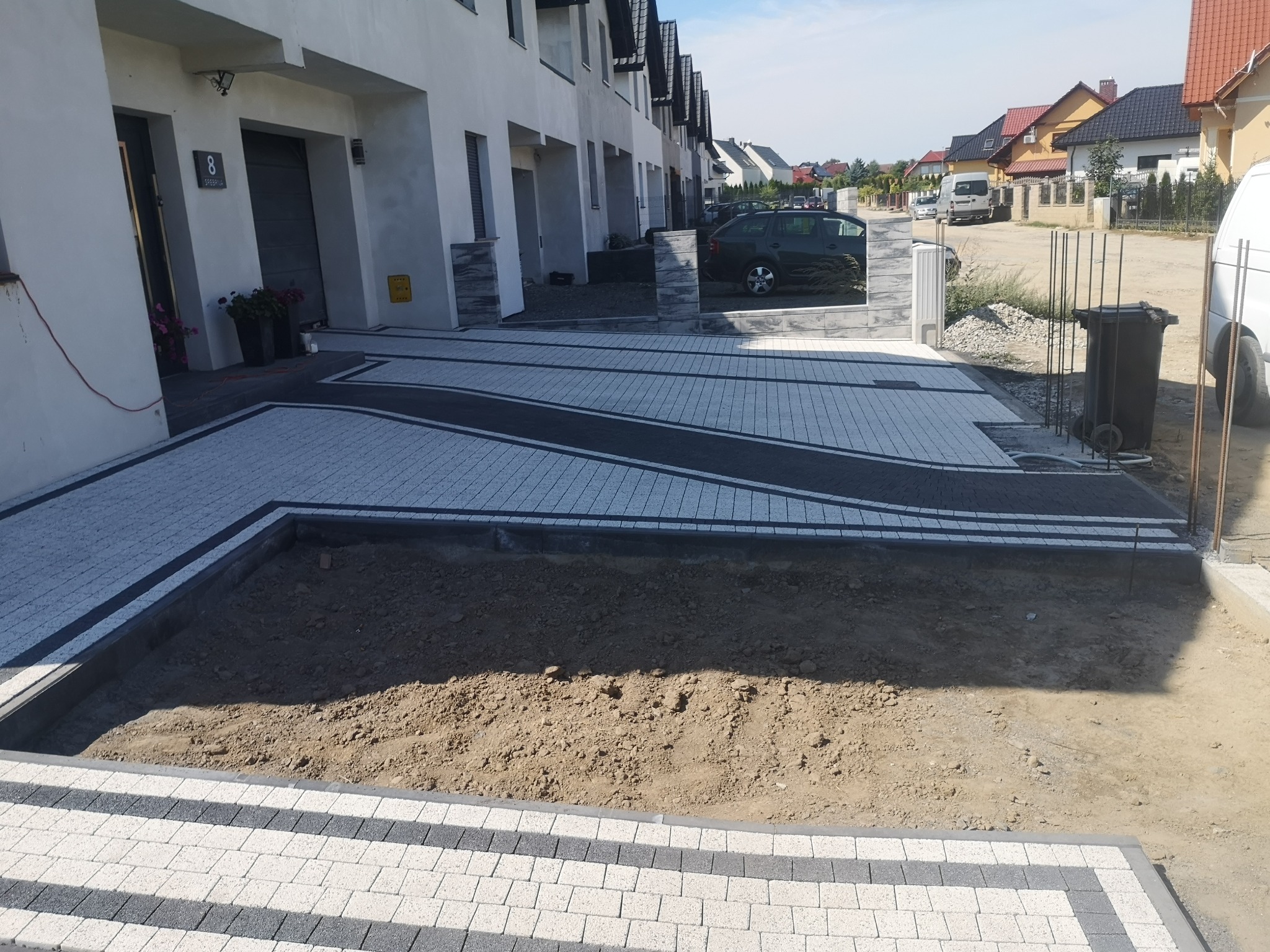 Nowoczesny podjazd z kostki brukowej w odcieniach szarości i bieli, z czarnymi akcentami, prowadzący do szeregu domów jednorodzinnych; w tle widoczne samochody i inne budynki, a w pierwszym planie...