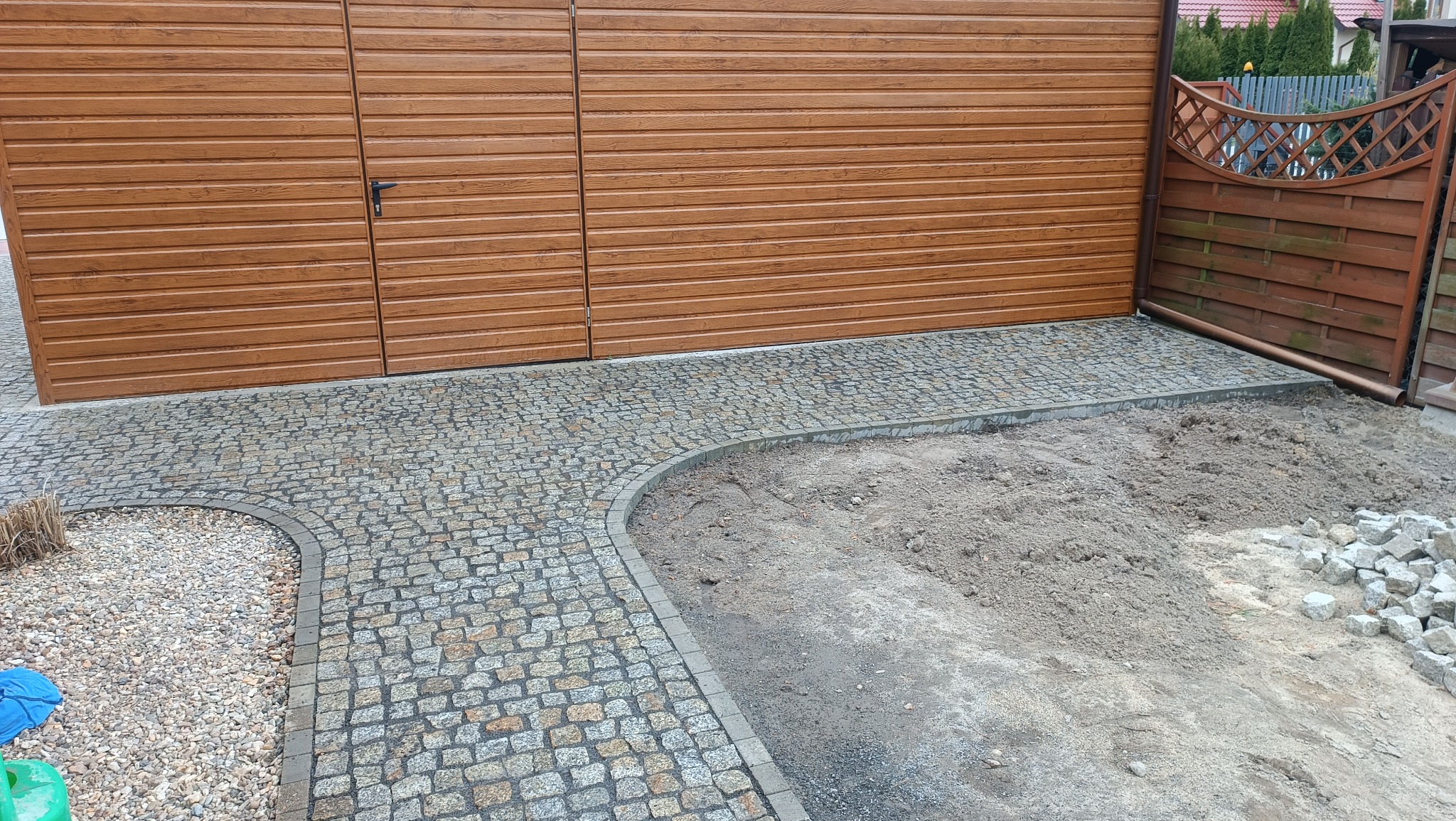 Ułożona kostka granitowa przed garażem z brązową bramą segmentową, widoczne obrzeża i przygotowany teren pod dalsze prace.