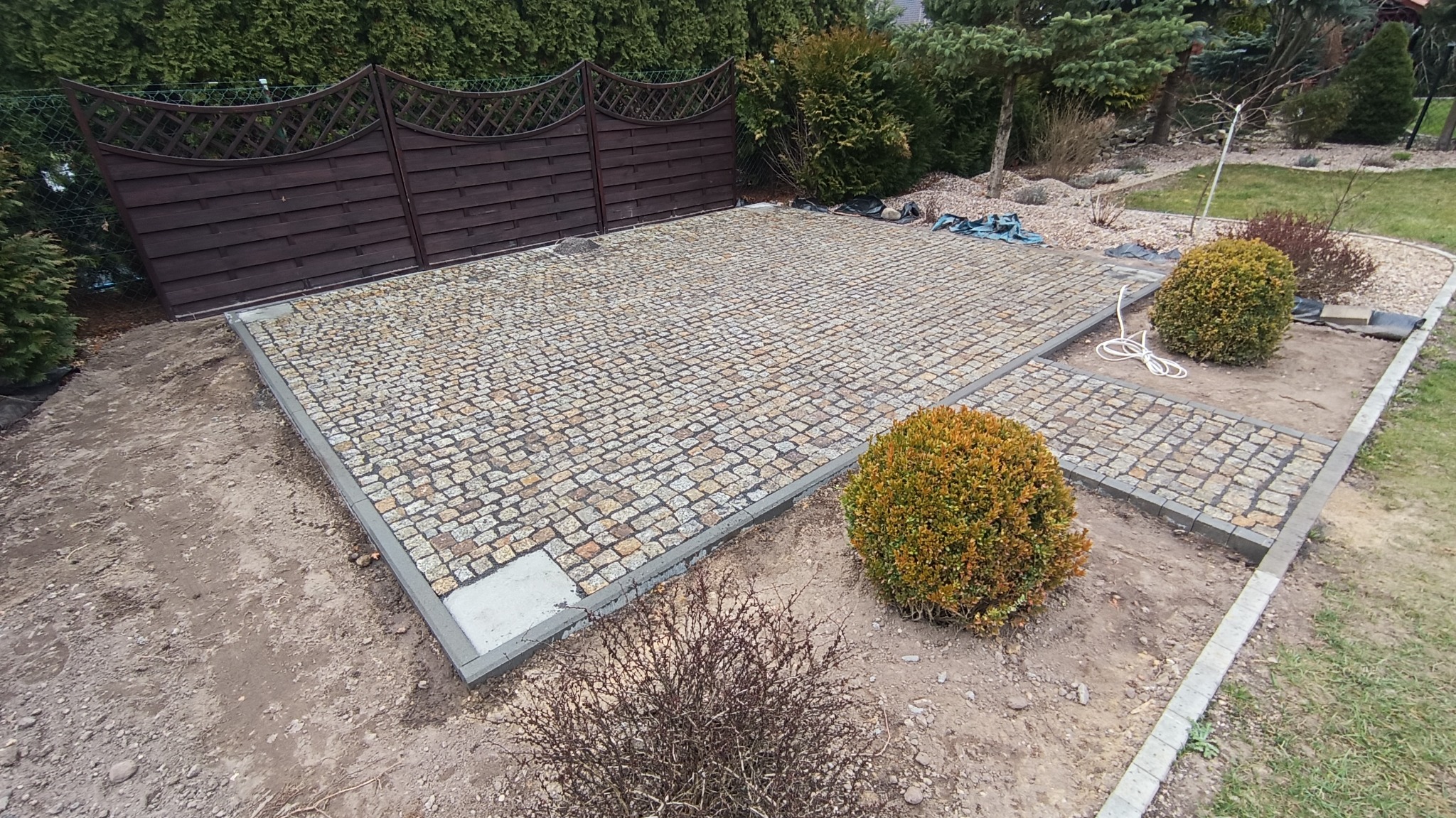Ułożona kostka brukowa w ogrodzie, widok z góry, z brązowym drewnianym płotem w tle, z dwoma okrągłymi krzewami i widocznym fragmentem trawnika. Kostka w odcieniach szarości i beżu, z szarą obwódką.