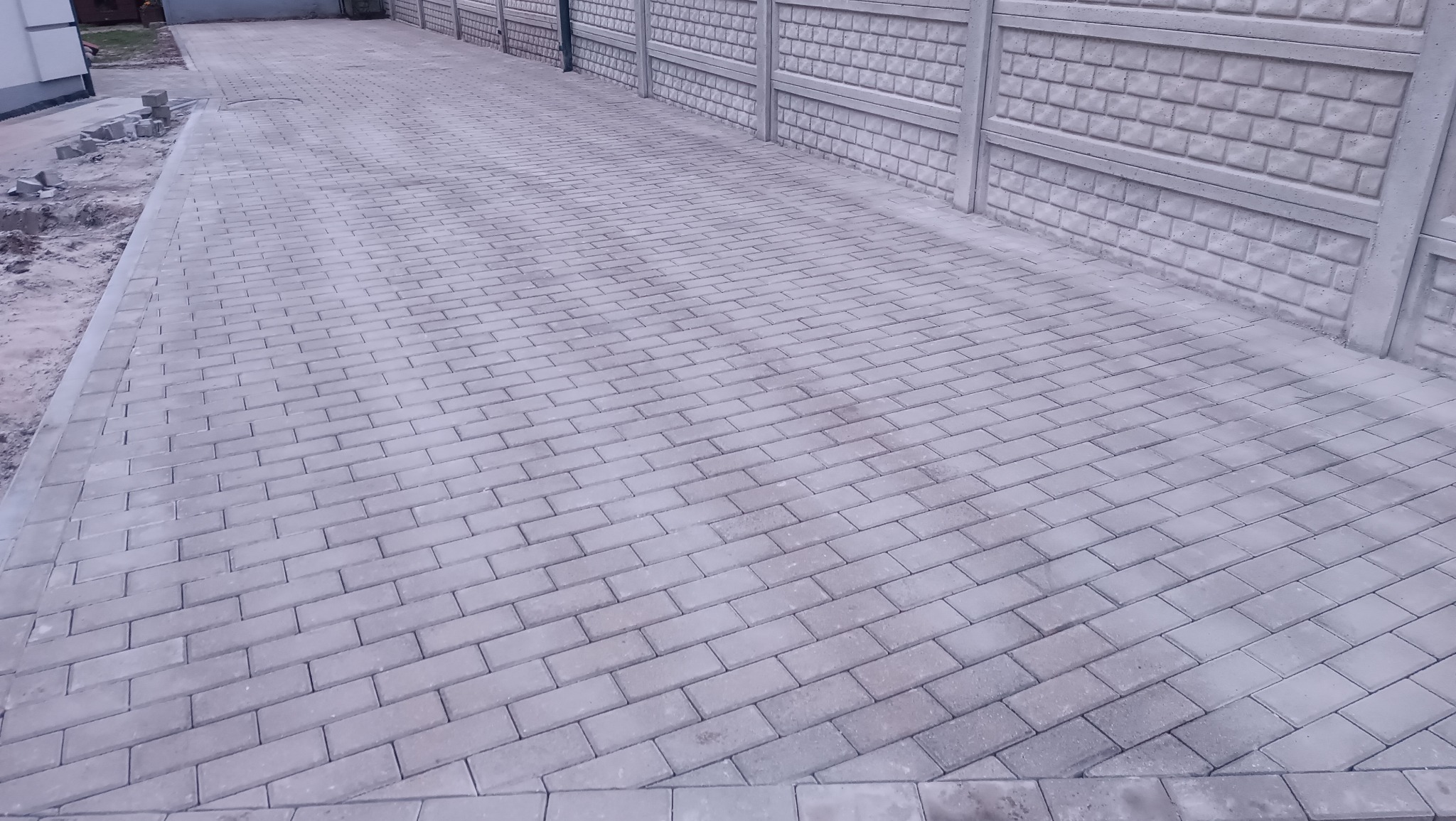 Świeżo ułożona, jasnoszara kostka brukowa tworząca podjazd, obramowana kostką brzegową. W tle betonowe ogrodzenie z fakturą cegły.