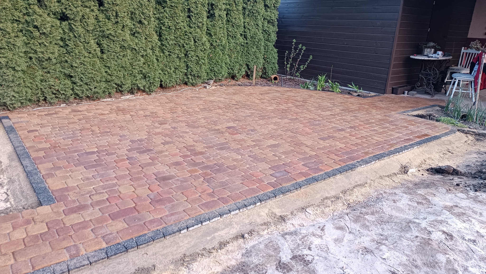 Nowo ułożona, czerwono-brązowa kostka brukowa z ciemnoszarą obwódką, widoczna na tle zielonego żywopłotu i ciemnego budynku, z fragmentami piasku i ziemi wzdłuż krawędzi.