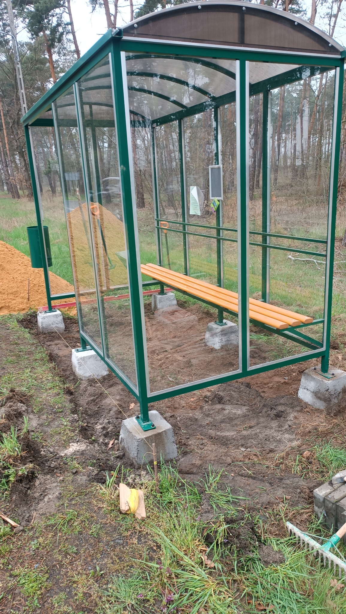 Zadaszona wiata przystankowa z zieloną konstrukcją i drewnianą ławką, osadzona na betonowych fundamentach, obok widoczny kosz na śmieci i przygotowany piasek; teren wokół wyrównany, widoczne...