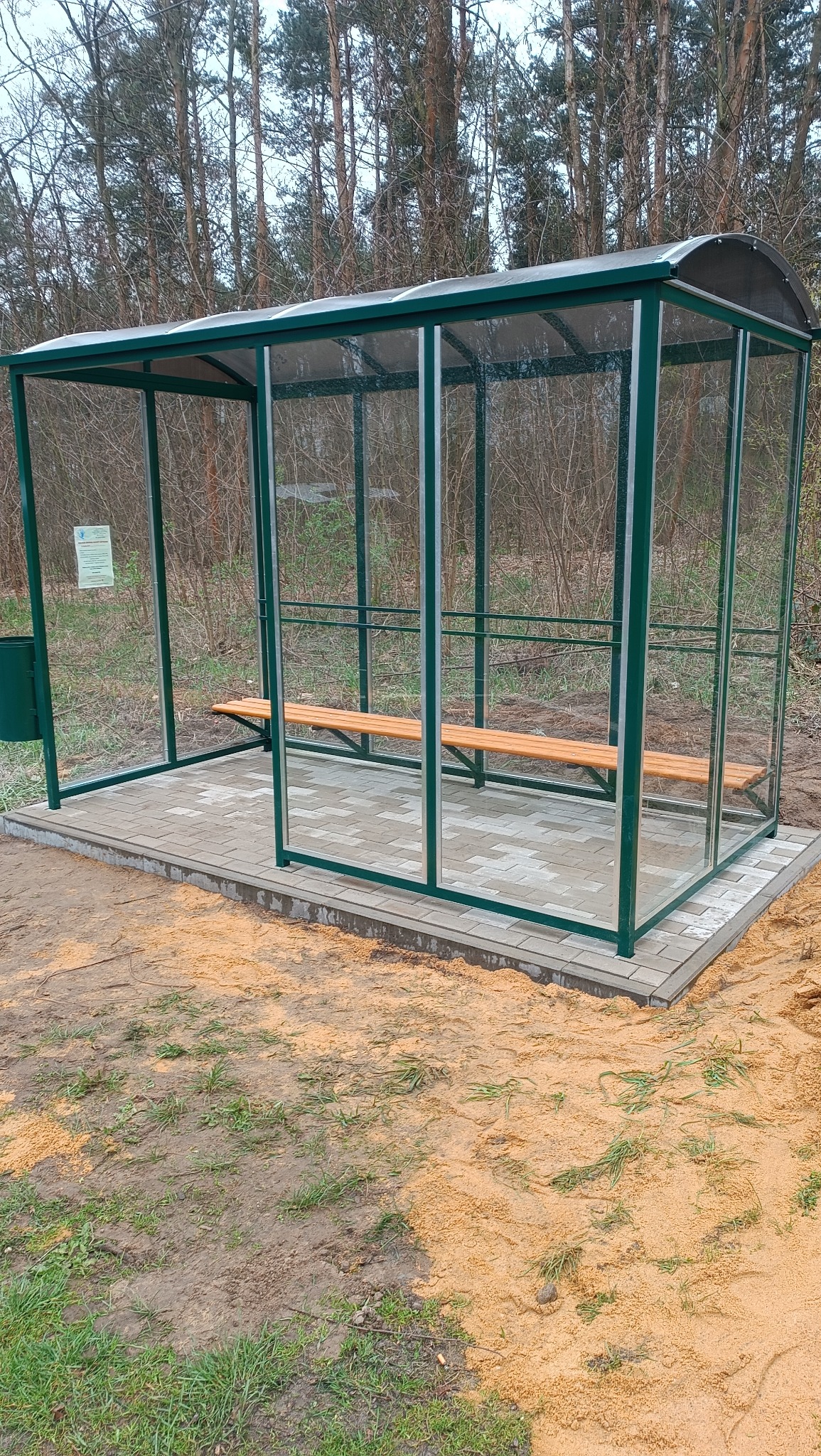 Nowoczesna, zielona wiata przystankowa z drewnianą ławką, osadzona na kostce brukowej, otoczona częściowo piaskiem i zielenią, z lasem w tle.