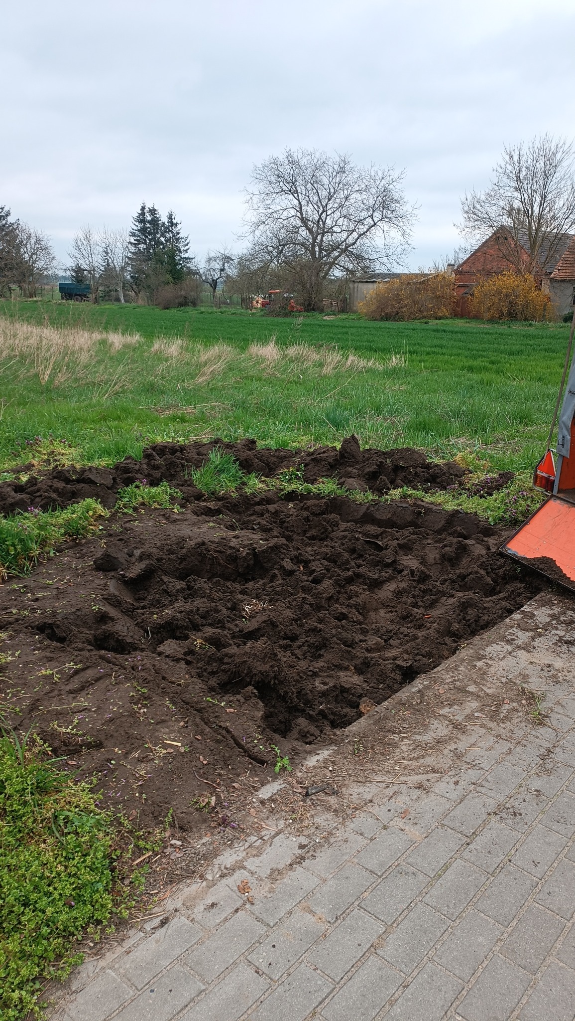 Świeżo przekopana ziemia na działce przylegającej do brukowanej ścieżki, na tle zielonej trawy i drzew, widoczny fragment pomarańczowego urządzenia.