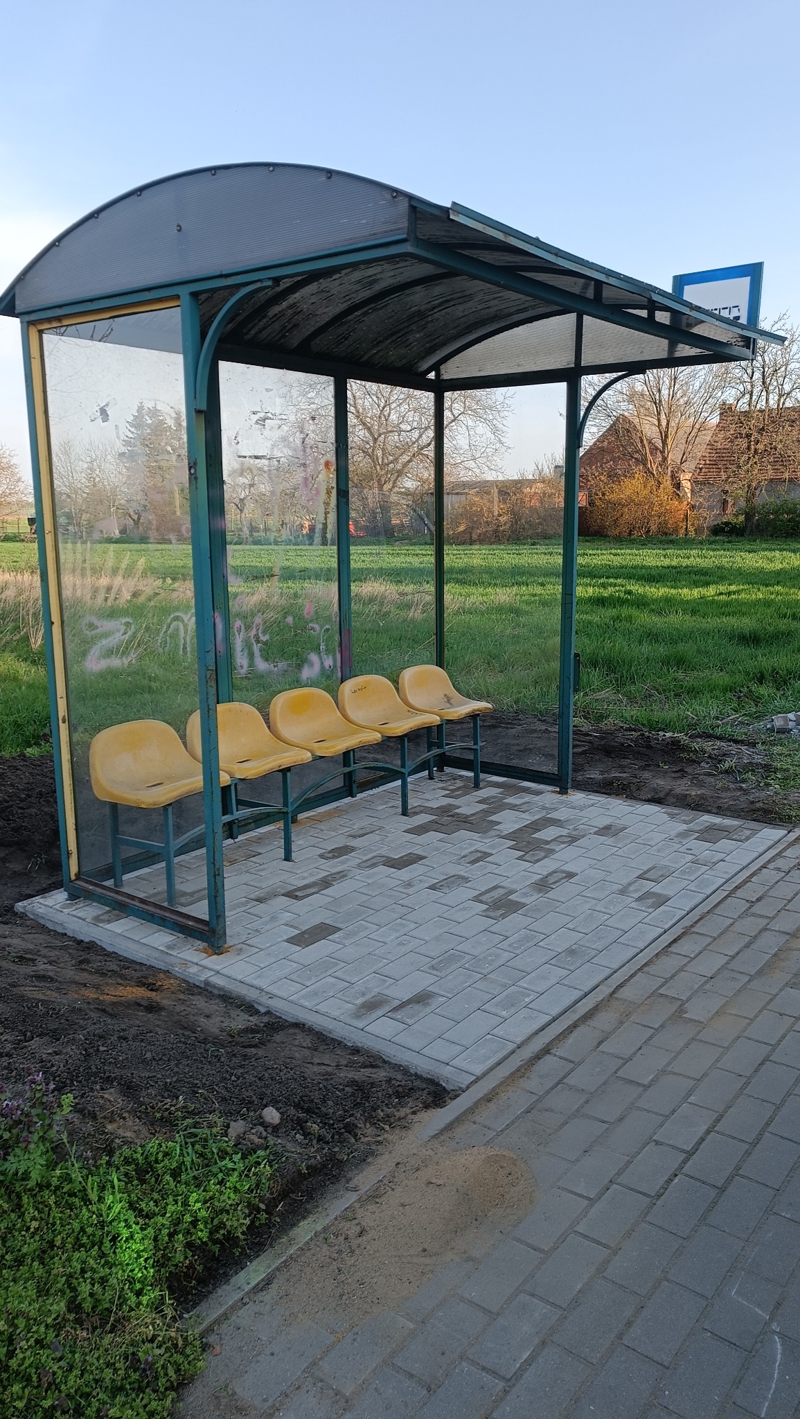 Nowo wybrukowana zatoczka autobusowa z żółtymi siedzeniami i przeszklonymi ścianami w wiejskim otoczeniu.