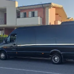Czarny, wysoki bus marki Mercedes-Benz Sprinter z przyciemnianymi szybami, zaparkowany na ulicy przed nowoczesnym domem z balkonem i palmą w tle.