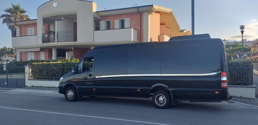 Czarny, wysoki bus marki Mercedes-Benz Sprinter z przyciemnianymi szybami, zaparkowany na ulicy przed nowoczesnym domem z balkonem i palmą w tle.