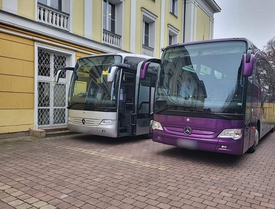 Dwa autobusy Mercedes: srebrny z otwartymi drzwiami i fioletowy, zaparkowane na brukowanym placu przed budynkiem z żółtą elewacją.