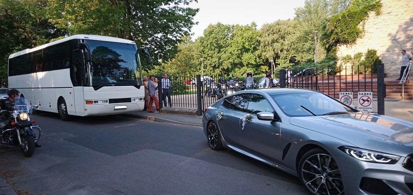 Biały autokar i szary samochód osobowy z dekoracjami ślubnymi stoją na ulicy, w tle grupa motocyklistów i goście weselni przed budynkiem z kamienia.