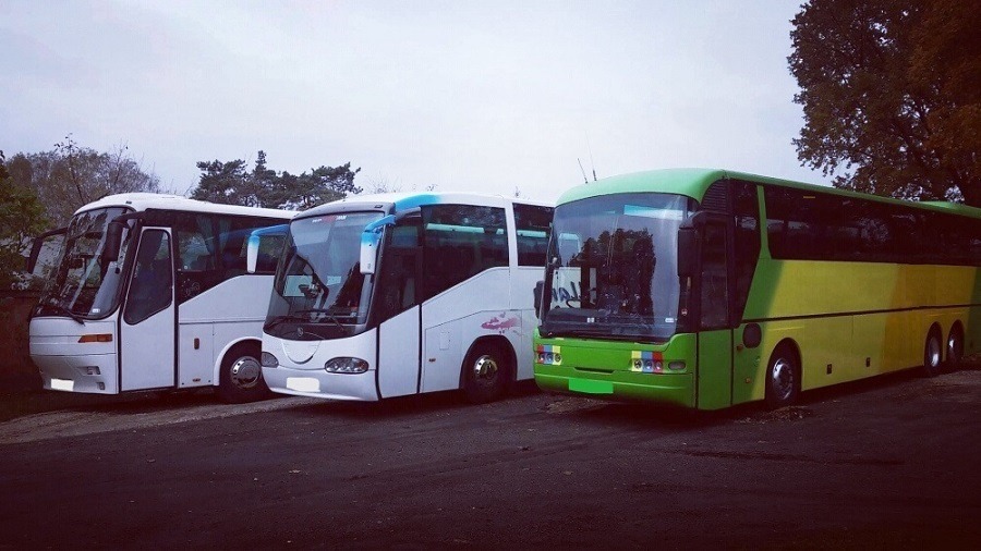 Trzy autokary turystyczne zaparkowane obok siebie: biały z otwartymi drzwiami, biały z niebieskimi akcentami i zielono-żółty, na tle drzew i pochmurnego nieba.