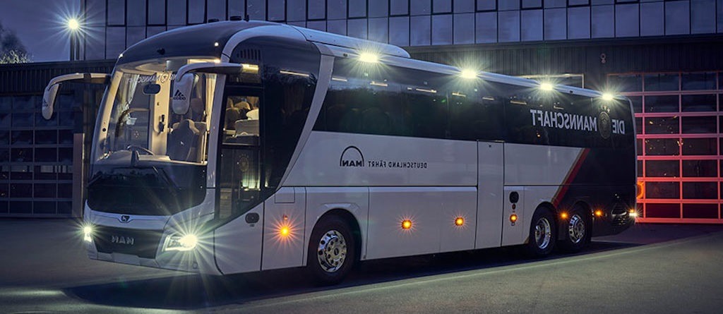 Nowoczesny autokar MAN w barwach biało-czarnych z czerwonym akcentem, oświetlony reflektorami, zaparkowany przed budynkiem z bramą garażową w nocnej scenerii.