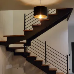Nowoczesne schody z drewnianymi stopniami i czarną metalową konstrukcją, oświetlone wiszącą lampą z czarnym abażurem, częściowo zabezpieczone folią ochronną.