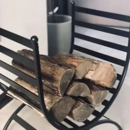 Metalowy stojak na drewno kominkowe w kształcie półksiężyca, wypełniony pociętymi kawałkami drewna, na tle jasnej podłogi i doniczki z rośliną.