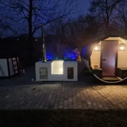 Oświetlona zewnętrzna sauna beczka i betonowa zabudowa z paleniskiem oraz jacuzzi z niebieskim podświetleniem, widoczne w nocy.