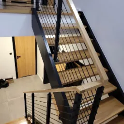 Nowoczesne schody z drewnianymi stopniami i czarnymi metalowymi elementami konstrukcyjnymi oraz balustradą z cienkich, poziomych prętów, widoczne z góry.