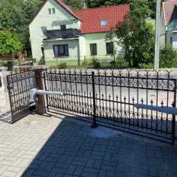 Otwarta, automatyczna brama wjazdowa z metalowymi zdobieniami, widoczny mechanizm otwierający, kostka brukowa na podjeździe, dom w tle.