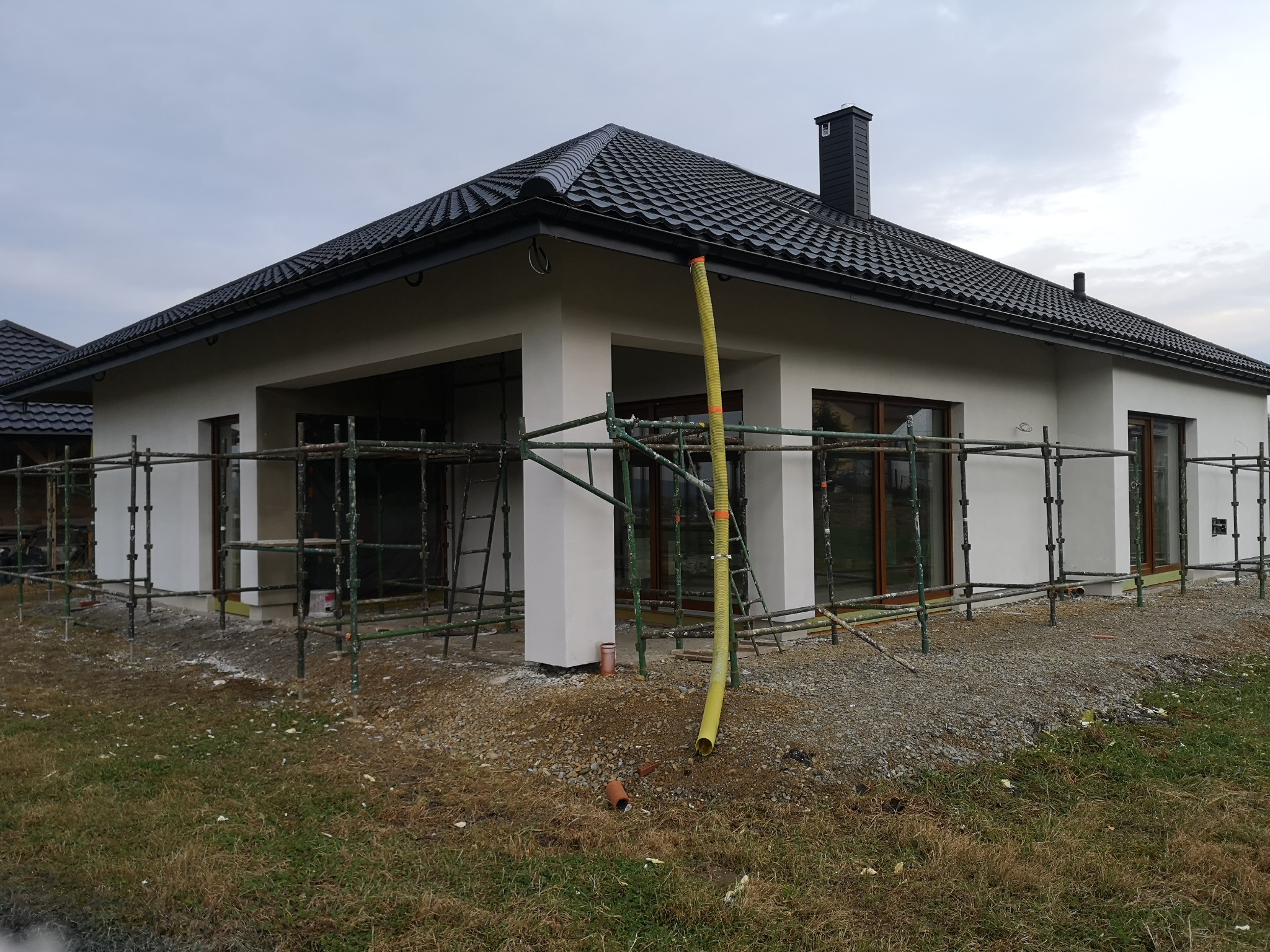 Nowoczesny dom jednorodzinny w trakcie prac elewacyjnych, widoczne rusztowania i żółty wąż wentylacyjny, biała elewacja, czarny dach z dachówki ceramicznej.