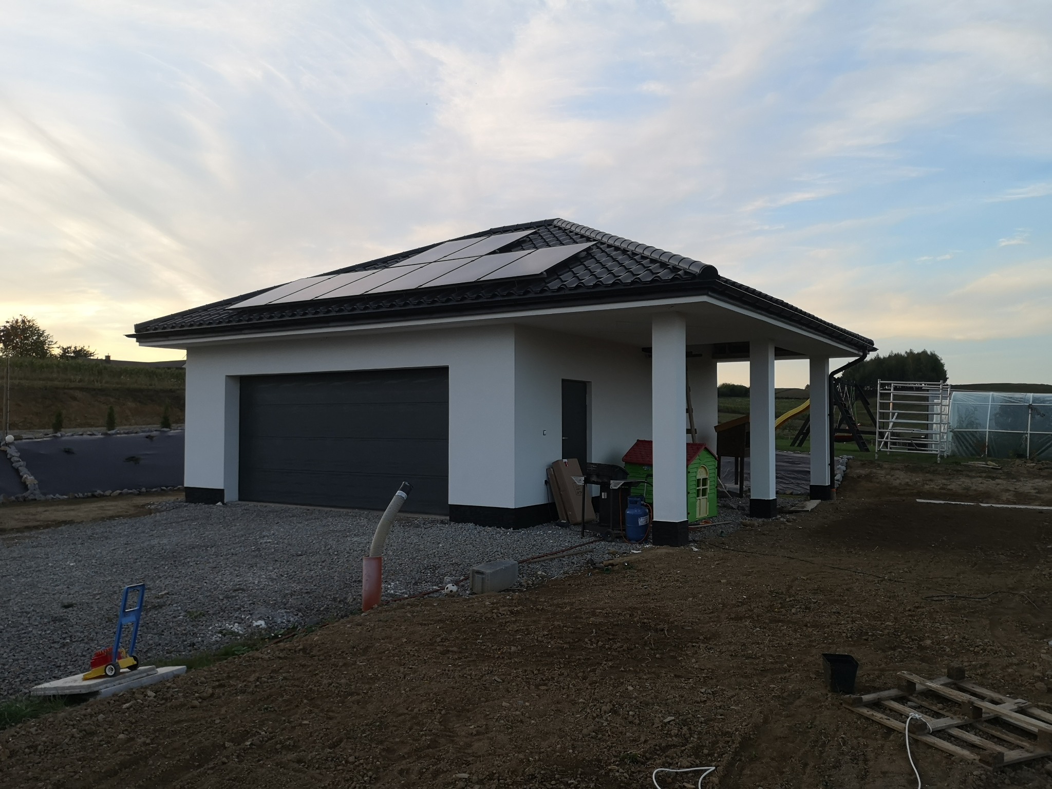 Nowoczesny parterowy garaż z panelami słonecznymi na dachu, szara brama garażowa, biała elewacja, otoczenie w trakcie zagospodarowywania, widoczny plac zabaw w tle.