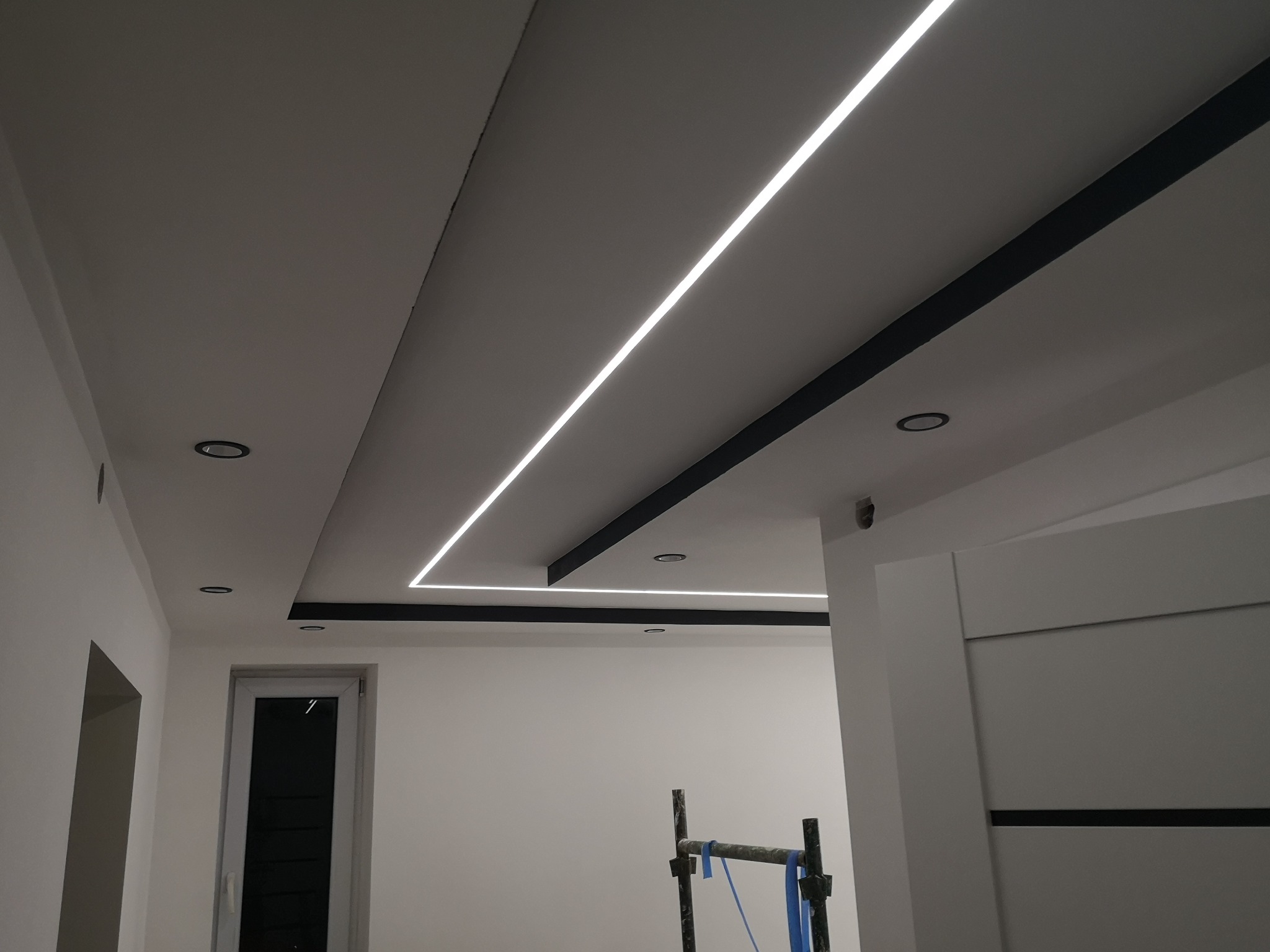 Nowoczesny sufit podwieszany z oświetleniem LED liniowym i czarnymi listwami dekoracyjnymi, widoczne również okrągłe lampy sufitowe i fragment rusztowania malarskiego.