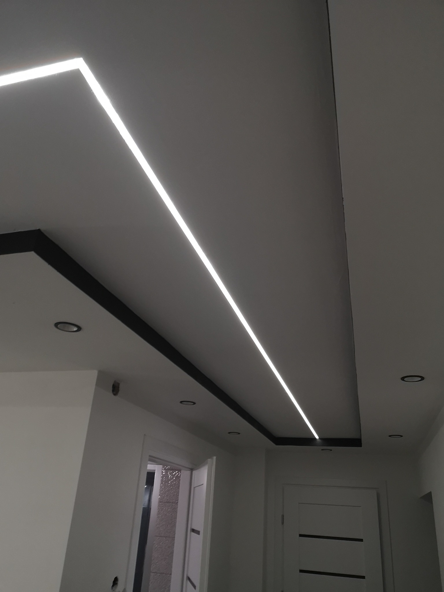 Nowoczesne oświetlenie sufitowe w korytarzu z podwieszanym sufitem i liniowymi lampami LED, widoczne drzwi wejściowe.