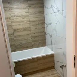 Wykończona łazienka z wanną obudowaną panelami drewnopodobnymi, płytkami drewnopodobnymi na ścianie i podłodze oraz białymi płytkami imitującymi marmur na ścianie, widoczne krzyżyki dystansowe...