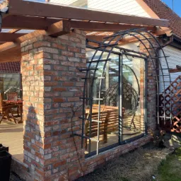 Murowana altana z cegły z drewnianą konstrukcją dachu, widoczny stół z krzesłami wewnątrz, metalowa konstrukcja pod pnącza przy szklanej ścianie i wiszący fotel.