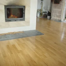 Jasna, lakierowana podłoga ułożona w jodełkę, widoczny kominek z paleniskiem i niedokończone obłożenie, listwy przypodłogowe przygotowane do montażu, w tle sprzęt remontowy i drabina.