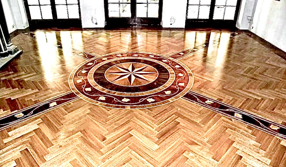 Wypolerowana podłoga z parkietu ułożonego w jodełkę z centralnym motywem rozety z gwiazdą, otoczona dekoracyjnym ornamentem roślinnym.