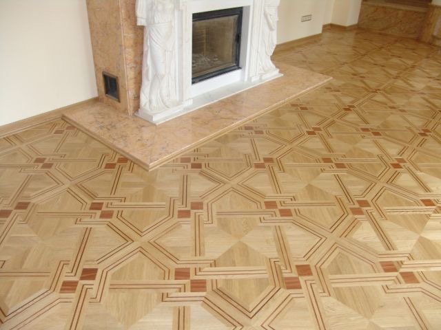 Wzorzysta drewniana podłoga ułożona w geometryczny deseń z ciemniejszymi kwadratami, sąsiadująca z marmurowym kominkiem z rzeźbami.