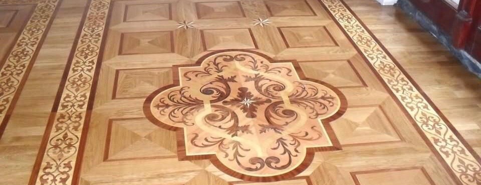 Drewniana podłoga z dekoracyjnym wzorem intarsji, centralny motyw floralny otoczony kwadratami i bordiurą z ornamentami.