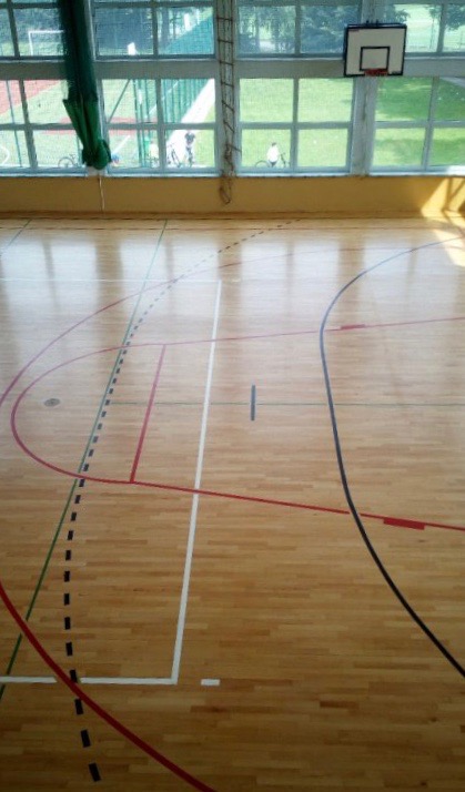 Parkiet w hali sportowej z liniami boiska do koszykówki, siatkówki i piłki ręcznej, widok na zewnątrz przez okna z siatką ochronną.