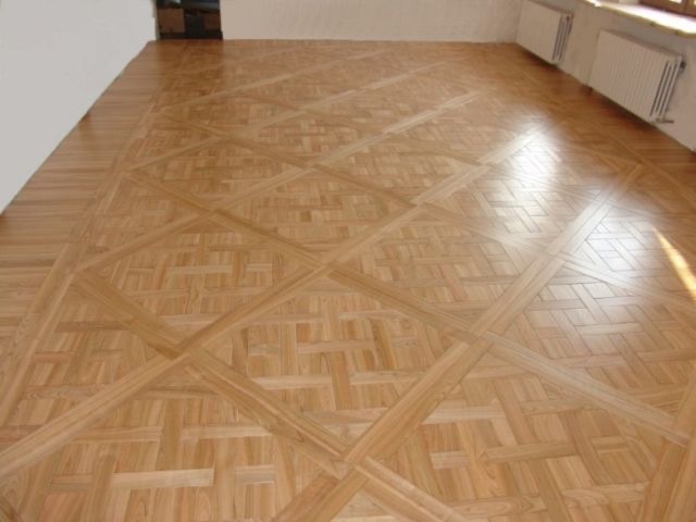 Jasny parkiet ułożony we wzór kwadratów z wypełnieniem w formie plecionki, widoczne fragmenty ścian i grzejniki