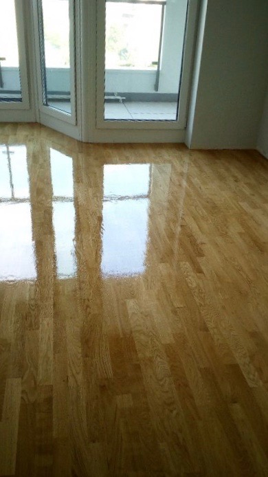 Lakierowana podłoga parkietowa z widocznymi refleksami światła z okien balkonowych, ukazująca gładką powierzchnię i naturalny rysunek drewna.