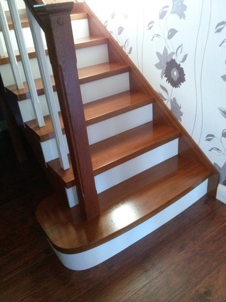 Odnowione drewniane schody z białymi podstopnicami i ciemnobrązową balustradą, widoczne na tle ściany z tapetą w kwiaty, z parkietem na podłodze.