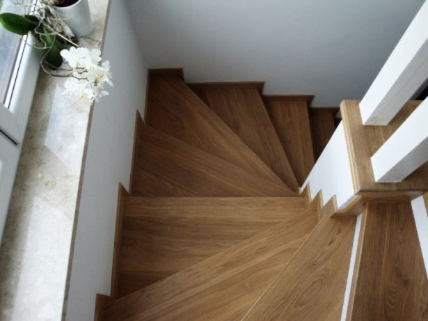 Drewniane schody z dębowymi stopniami, białe ściany i balustrada, widok z góry, obok okna stoi doniczka z białym storczykiem.