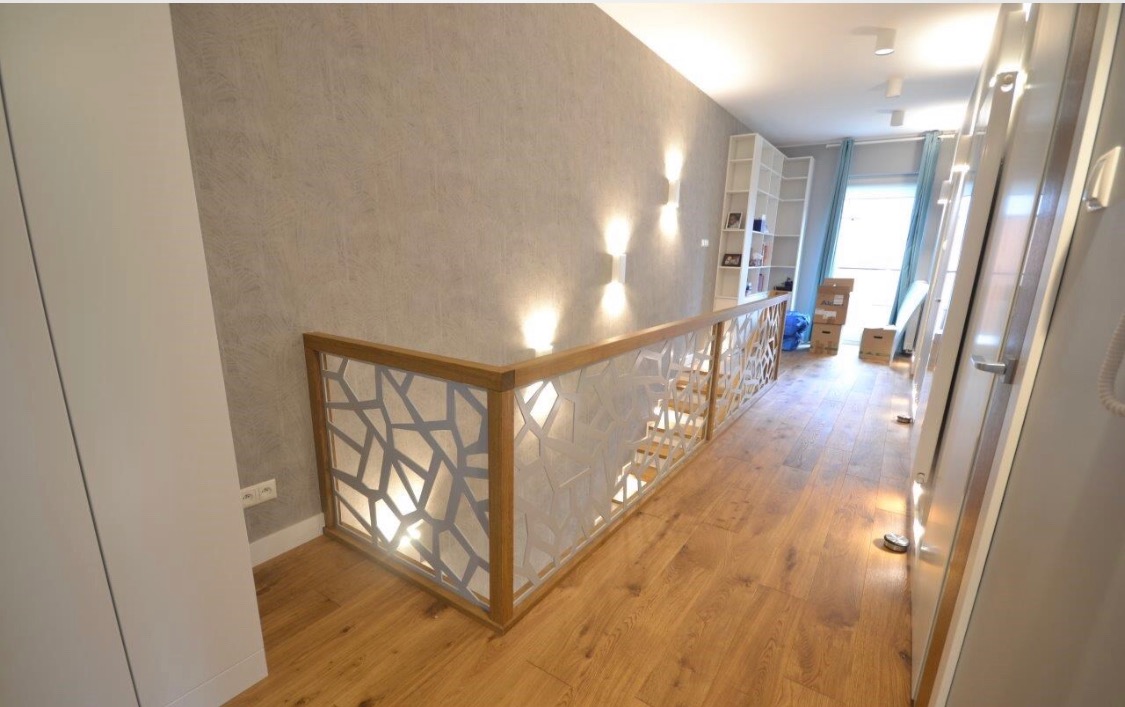 Nowoczesna balustrada schodowa z drewnianą ramą i ażurowym wypełnieniem w geometryczne wzory, parkiet na podłodze, szara ściana z kinkietami, widok na korytarz z regałem i kartonami.