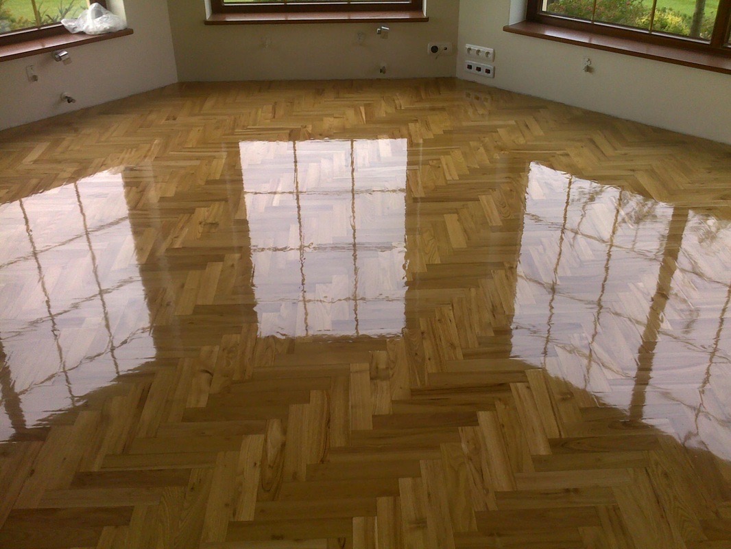 Błyszcząca, świeżo polakierowana podłoga z parkietu ułożonego we wzór jodełki, odbijająca okienne ramy i światło, w jasnym pomieszczeniu z białymi ścianami.