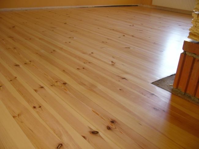 Jasna, drewniana podłoga ułożona z desek, widoczne sęki i naturalne usłojenie, fragment ceglanego kominka w rogu pomieszczenia.