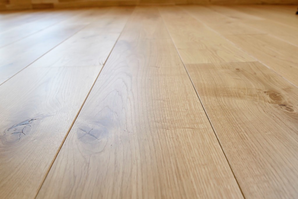 Jasna, drewniana podłoga z widocznymi słojami i naturalnymi sękami, ułożona z desek z minimalnymi szczelinami, sfotografowana z niskiego kąta, ukazująca perspektywę i głębię pomieszczenia.