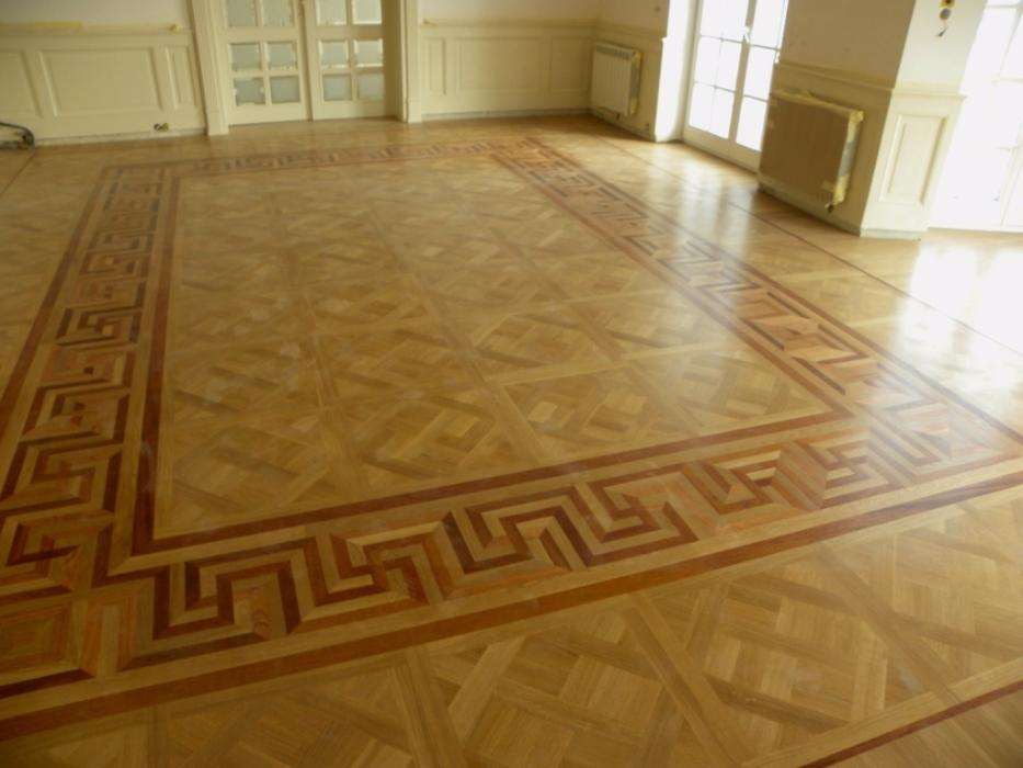 Parkiet ułożony we wzór geometryczny z bordiurą w stylu greckim, widok na duży salon z białymi ścianami i drzwiami balkonowymi.