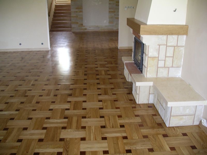 Parkiet ułożony we wzór koszykowy, z jasnego drewna przeplatanego ciemniejszymi elementami, w przestronnym salonie z kominkiem obłożonym jasnym kamieniem i schodami w tle.